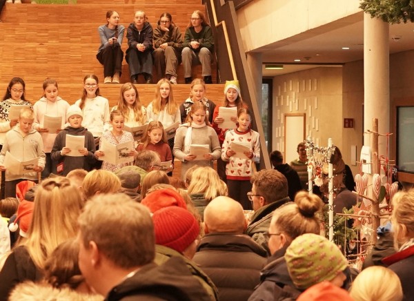 Ausschnitt der Pausenhalle bei der Eröffnung des Basars: Auf den untersten Stufen der Freitreppe singt der Unterstufenchor; im Vordergrund dichtes Gedränge, rechts ein Verkaufsstand mit Weihnachtsdeko.
