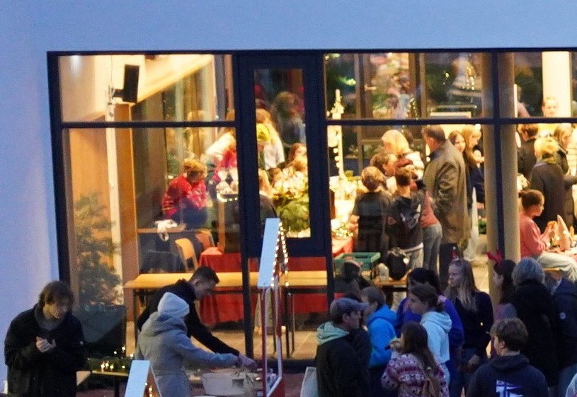 Blick aus der Vogelperspektive, der einen Ausschnitt des Innenhofs mit Essensverkauf zeigt und vielen Menschen, die sich angeregt unterhalten. Im Hintergrund ist die hell erleuchtete Pausenhalle zu erkennen, in der sich die Verkaufsstände der Klassen befinden